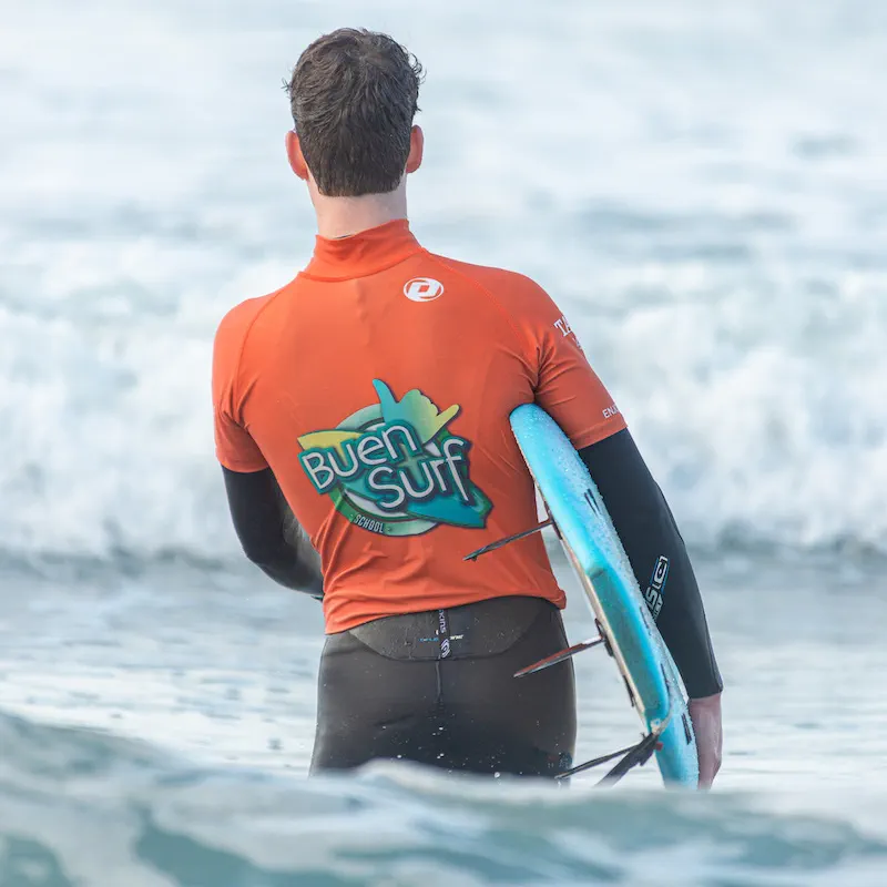 A surfer in a Buen Surf jersey wading out with a board under one arm